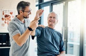 An elderly man wearing a dark blue t-shirt smiles at the physical therapist wearing a gray polo and glasses as he holds the patient's arm up to check his range of motion.