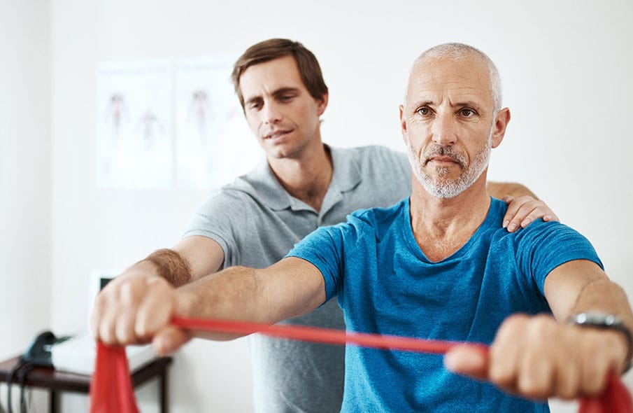 An older man with white stubble stares ahead in concentration while wearing a blue t-shirt and a black watch while holding a red exercise band in his outstretched hands as a physical therapist in a gray polo holds onto the band with one hand and the man's shoulder with the other.