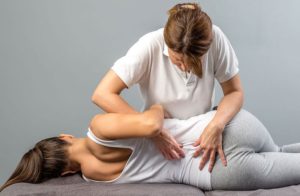 A brunette woman lays on her left side on a table with her back toward the camera. Her hair is pulled back in a pony tail and she wears a white tank top and light gray leggings. A medical professional woman with her brown hair pulled back in a pony tail leans over her to study the patient's lower back and presses her fingers on both sides of the patient's spine.